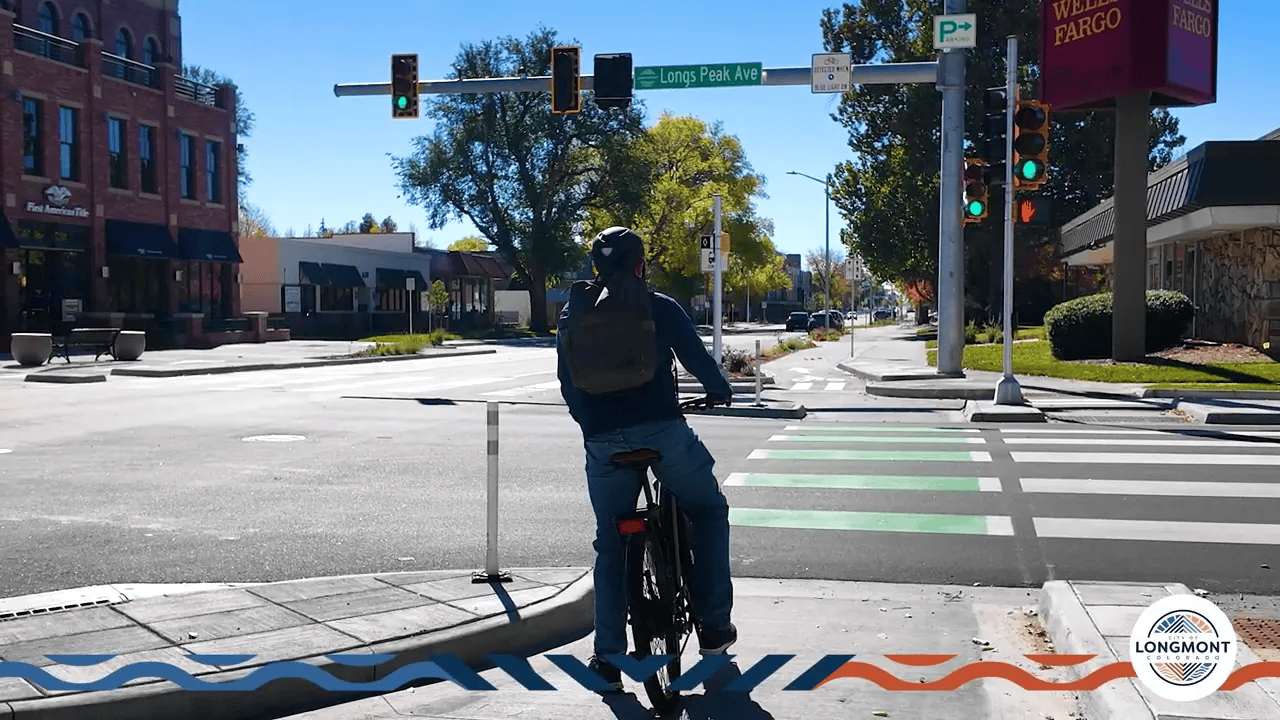 Cyclist stopped in a bike waiting area at a protected intersection with green bike crosswalk markings and traffic signals (Longs Peak Avenue).