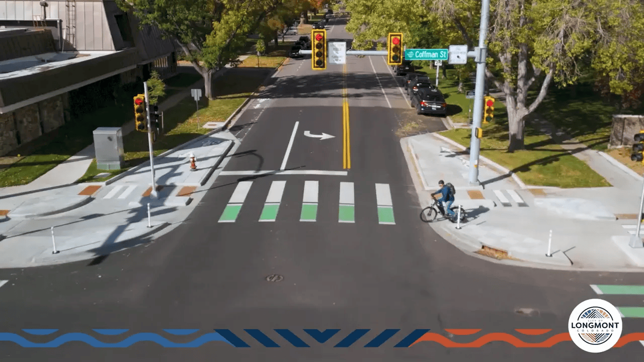 Aerial view of a protected intersection showing corner islands, green bike crosswalk panels, white pedestrian crosswalk, and a bicyclist staged in the bike waiting area.
