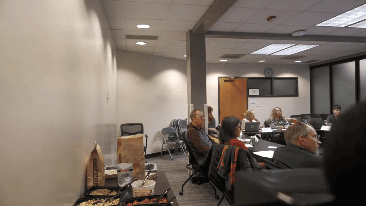 Clear wide shot of a commission meeting in a conference room with attendees seated around a table and a small buffet on a side table.