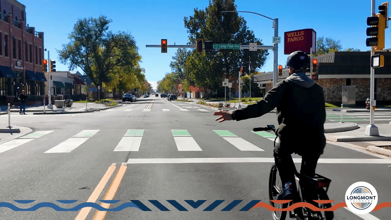 Street-level rear view of a cyclist approaching a protected intersection with green bike crossing panels and white pedestrian crosswalks.